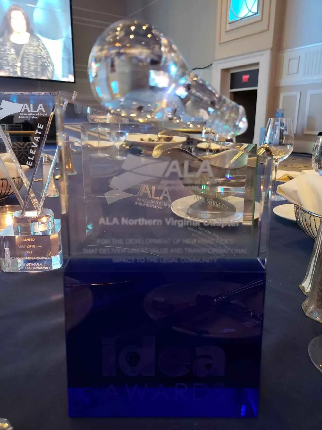 Transparent award plaque with engraved text and logo, placed on a table set for a formal event. The background shows glasses, a spoon, and a tray, with a TV screen and decorative wall elements.