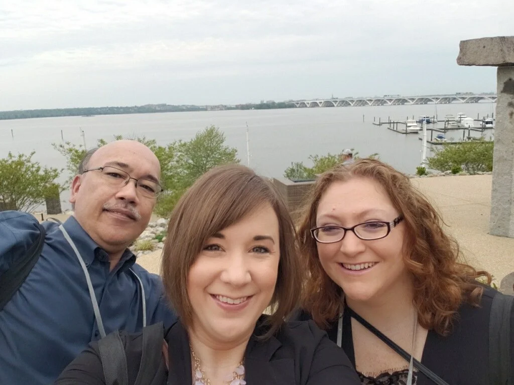 Three people taking a selfie outdoors near a body of water with boats and a bridge in the background.