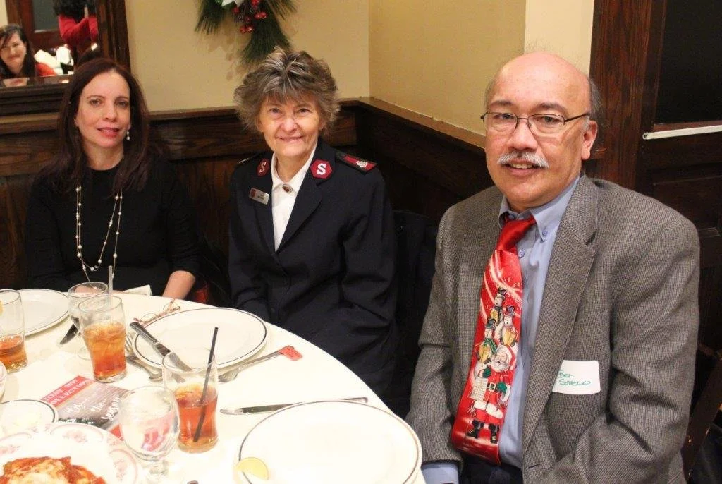 Three people sitting at a table in a restaurant, smiling at the camera. The woman on the left has long dark hair, the woman in the middle is wearing a uniform, and the man on the right is wearing glasses, a gray blazer, and a festive tie with holiday