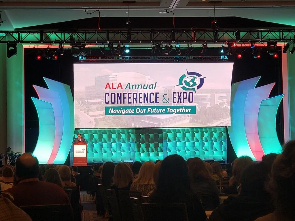 Audience attending the ALA Annual Conference and Expo with a large stage and a speaker at the podium. The screen behind displays the event logo and slogan, 'Navigate Our Future Together.'