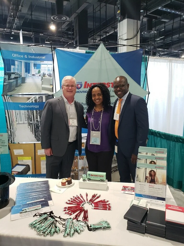 Three people standing behind a display table at a trade show booth, smiling, with promotional materials and promotional items on the table.