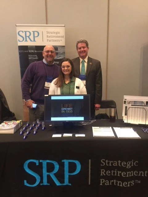 Three people standing behind a table at an event, with a sign that reads 'SRP Strategic Retirement Partners', and a computer monitor displaying 'LIFE EXPECTANCY IN 1935'.
