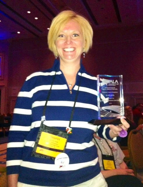 Smiling woman with blond hair holding a glass award in an indoor conference setting wearing a blue and white striped shirt and conference badge.