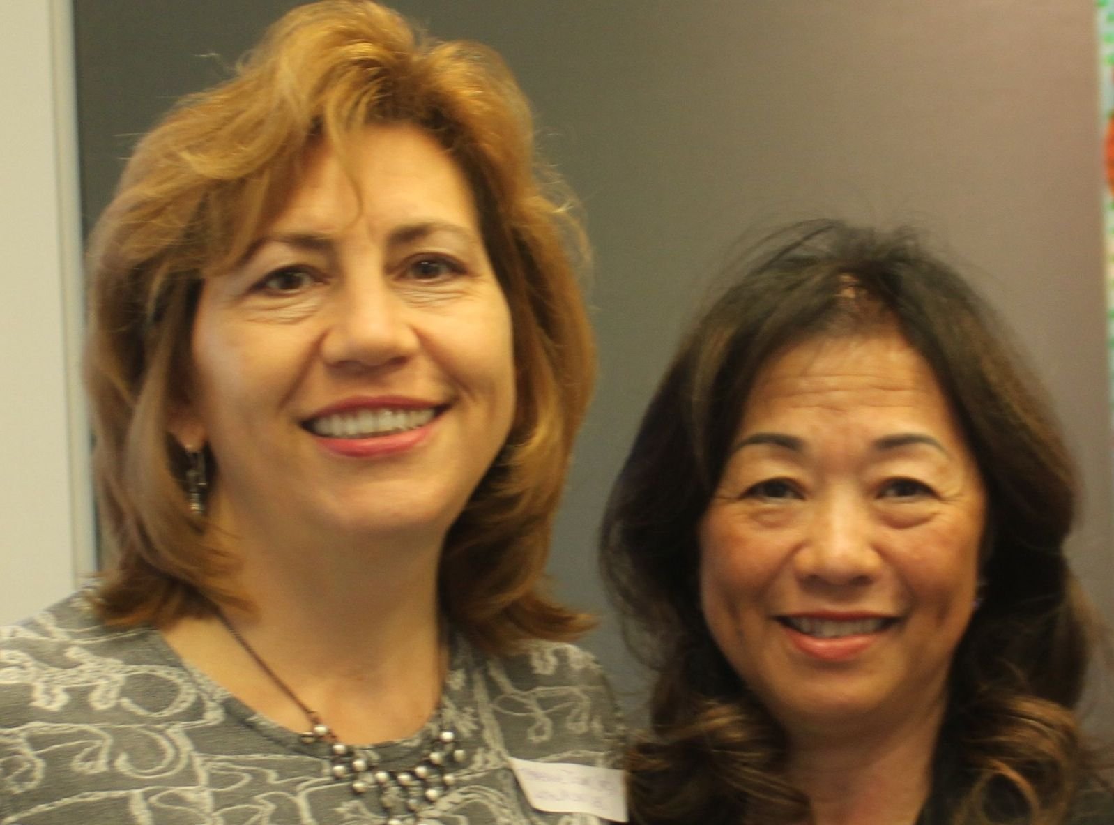 Two women smiling, standing close together indoors. One has shoulder-length light brown hair and is wearing a patterned top with a necklace. The other has shoulder-length dark hair and is wearing a dark top.