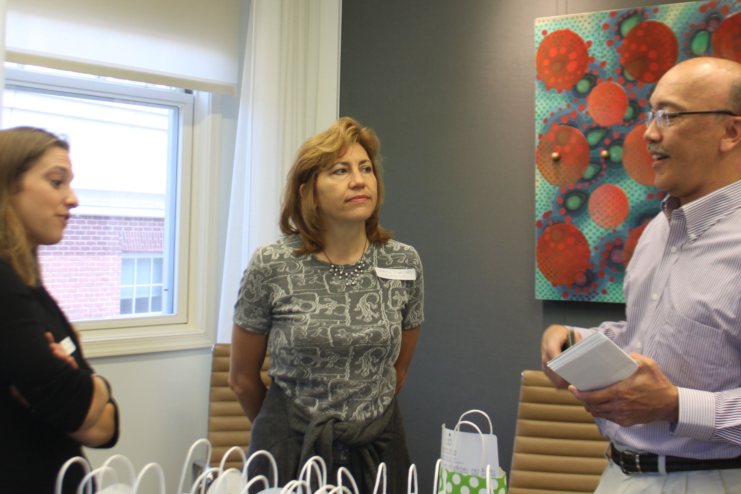 Three women and one man engage in conversation in a room with modern decor. The woman in the center has shoulder-length red hair and is wearing a patterned gray top. The woman on the left has light brown hair and is wearing a dark blazer. The woman o