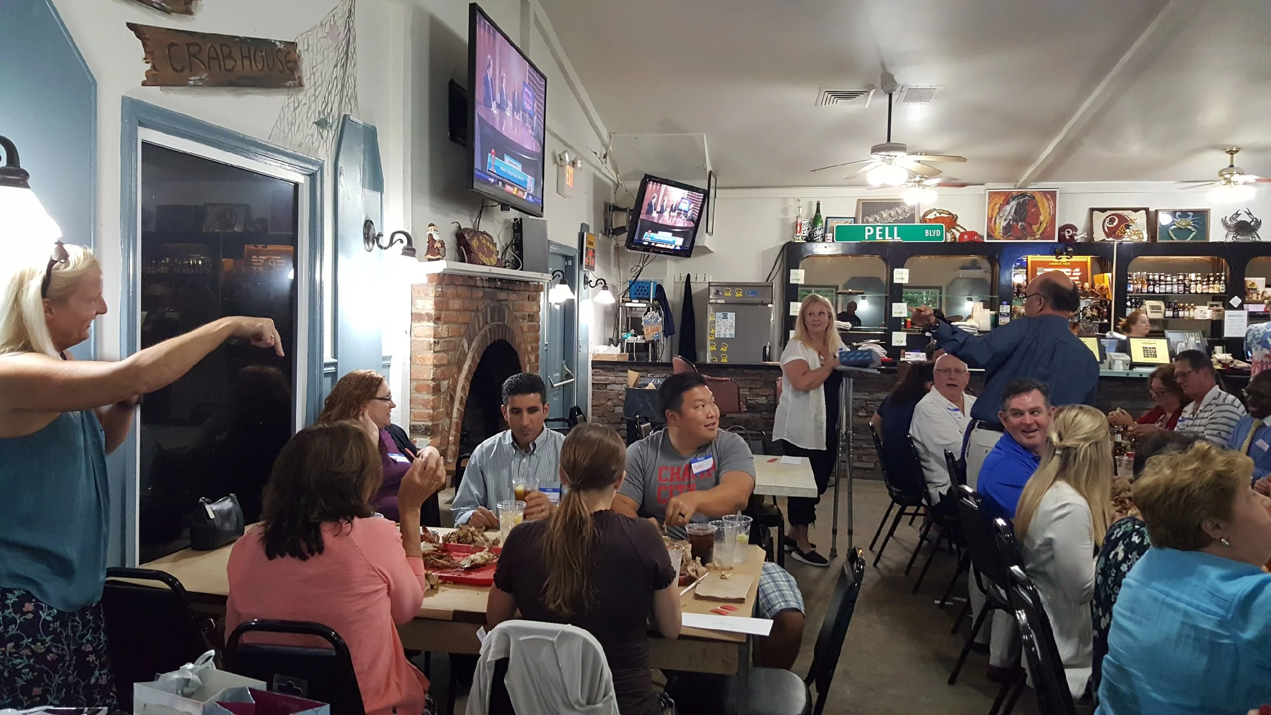 People gathered inside a restaurant, seated at tables, engaged in conversation and enjoying food and drinks. There are multiple TV screens mounted on the wall, and a woman is standing near the counter, while a man appears to be speaking to others. Th