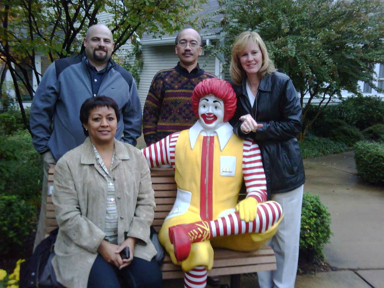 Four women and one man standing outdoors around a Ronald McDonald statue. Two women are seated, and two women and one man are standing behind the statue. There are bushes and trees in the background.