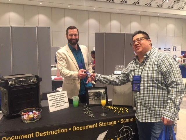 Two men at a trade show booth, one handing a raffle ticket to the other. The booth advertises document destruction, storage, and scanning services. One man is smiling, wearing a white blazer; the other, dressed casually, is also smiling. There is a f
