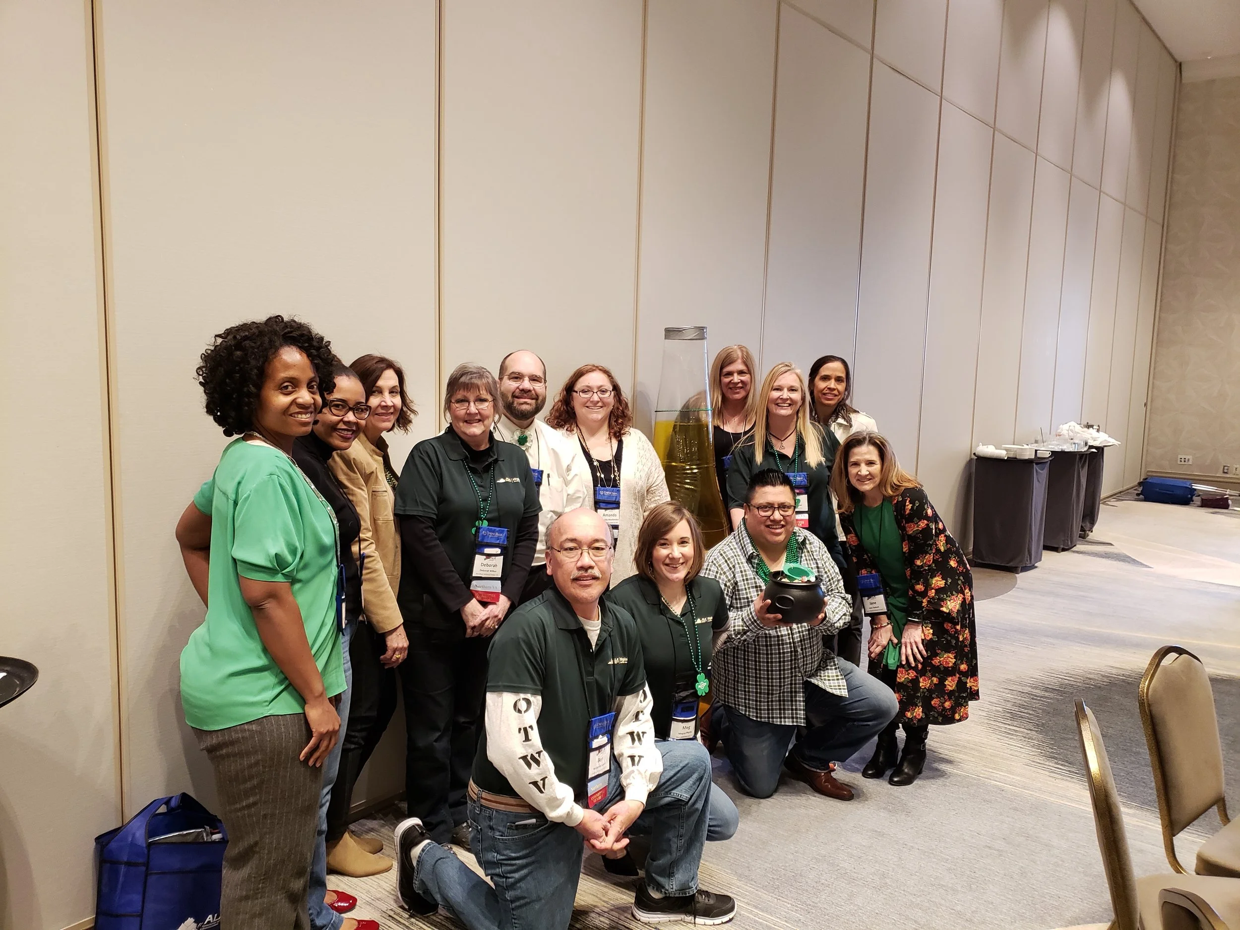 A group of diverse people smiling and posing together at an indoor event, with some wearing name tags and casual clothing, arranged in two rows in front of a plain wall.