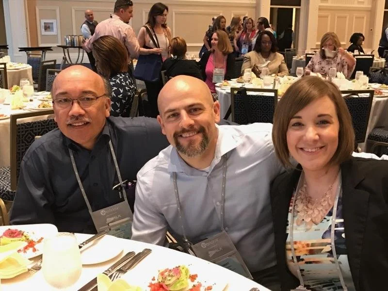 Three people smiling and sitting at a banquet table during a conference or event, with other attendees and banquet tables in the background.