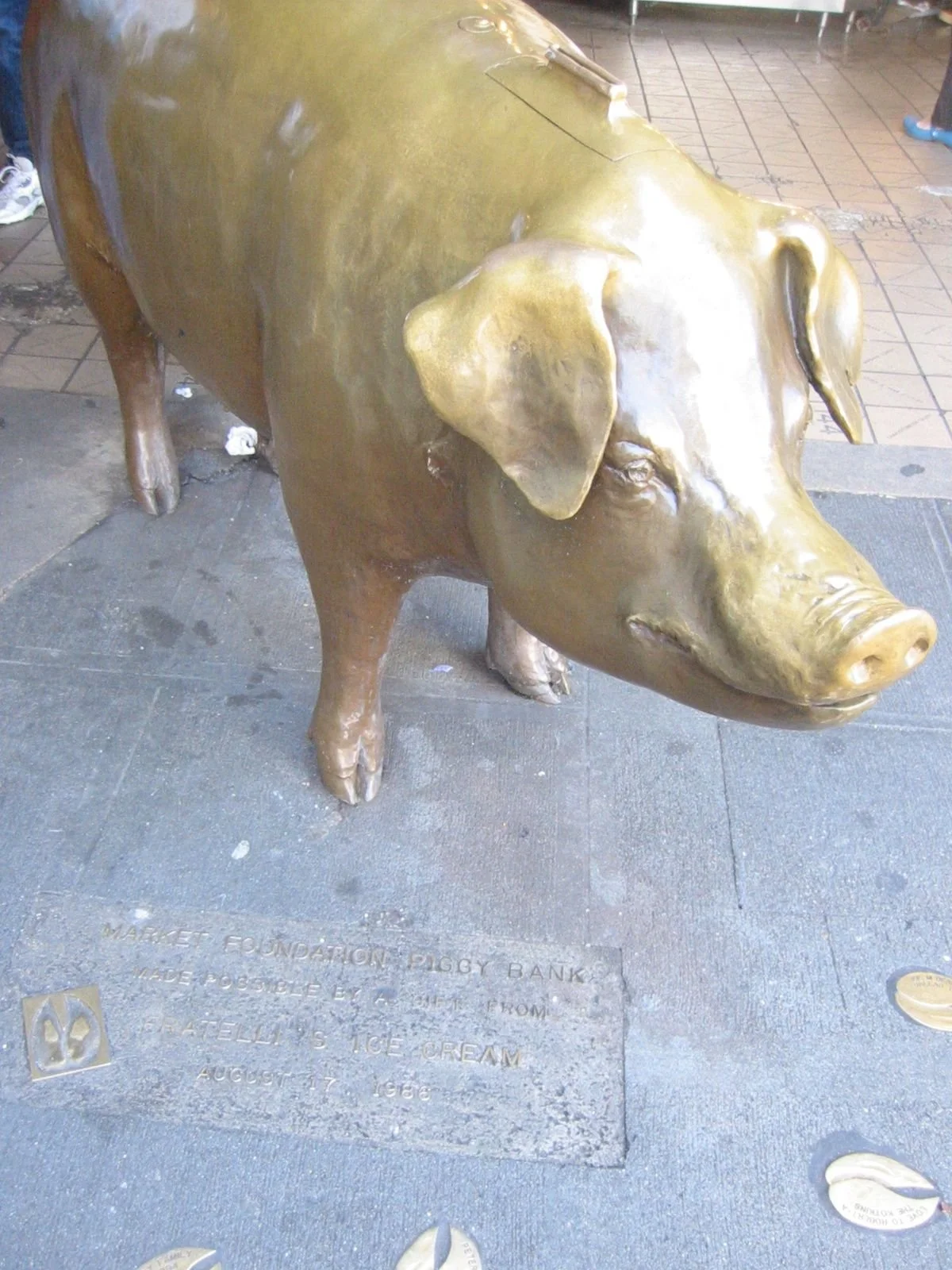 Bronze sculpture of a pig on a city sidewalk. The sculpture is located on a plaque that indicates it is a piggy bank made from a pig that was a foundation pig, created by Arlene L. Moe Cream in 1963.