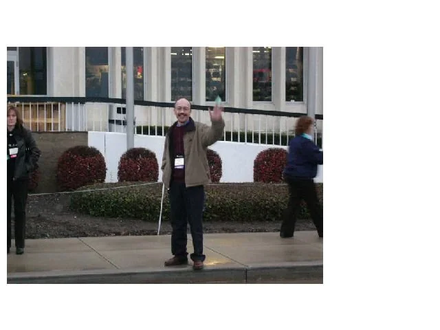 A man standing on a sidewalk waving with a big smile, wearing glasses, a brown jacket, and a dark shirt. Two women are walking in the background near a building with large windows.