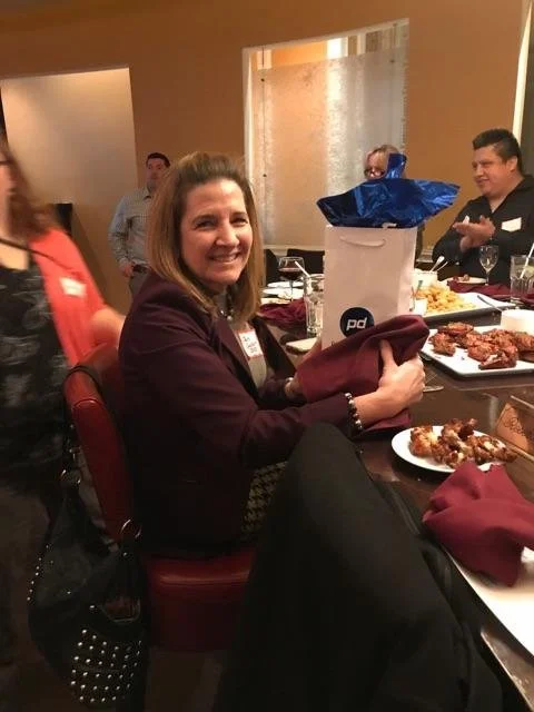 Woman smiling at a dinner table with food and a gift bag, surrounded by other guests, in a warmly lit restaurant or banquet setting.
