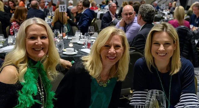 Three smiling women sitting at a table during a formal event or dinner, with a crowded banquet hall in the background.