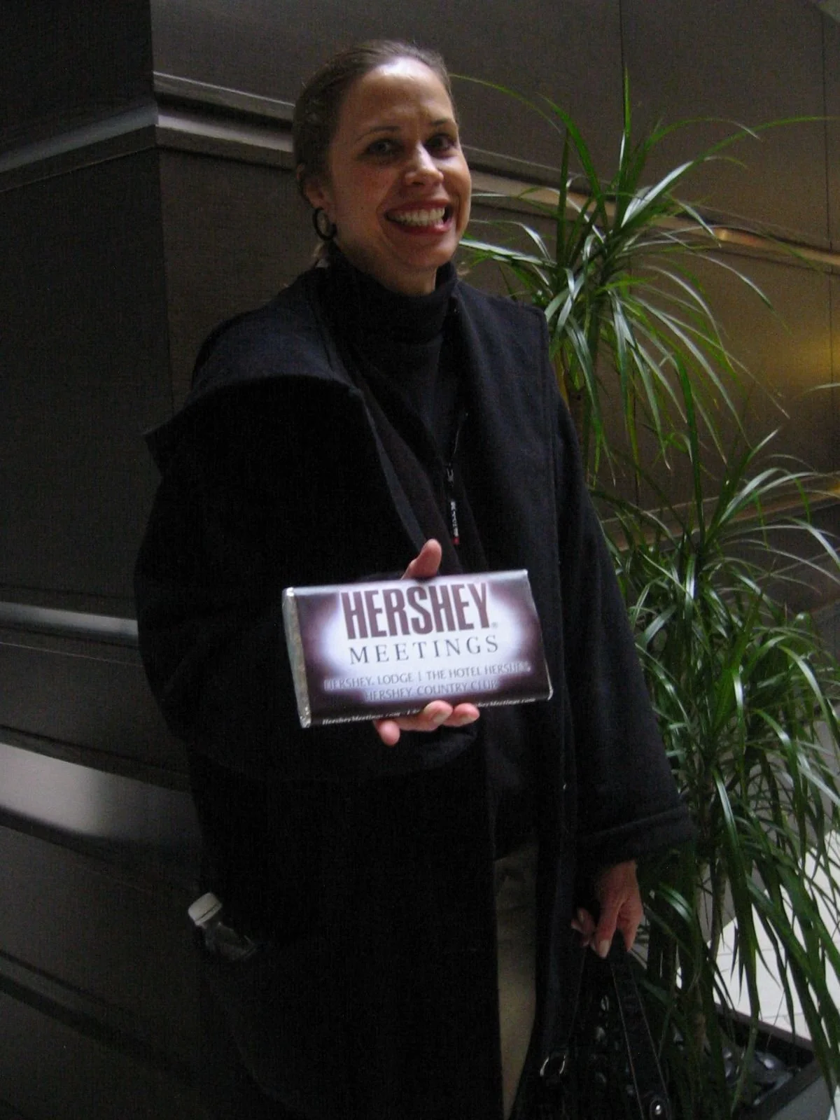 A woman smiling and holding a Hershey's chocolate bar with a label that reads "Hershey Meetings". She is standing next to a green plant and appears to be at an indoor event or meeting.