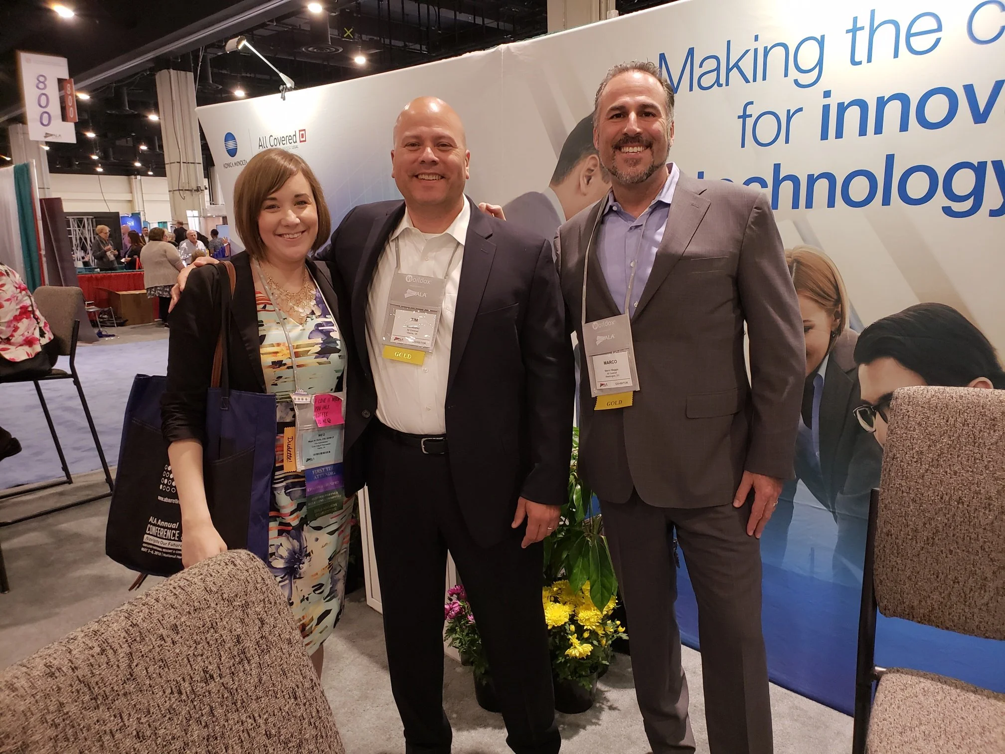 Three smiling people posing at a conference or expo, with two men wearing suits and a woman in a floral dress. They are standing in front of a large sign with blue text and a flower arrangement at the base. The background shows a busy event space wit