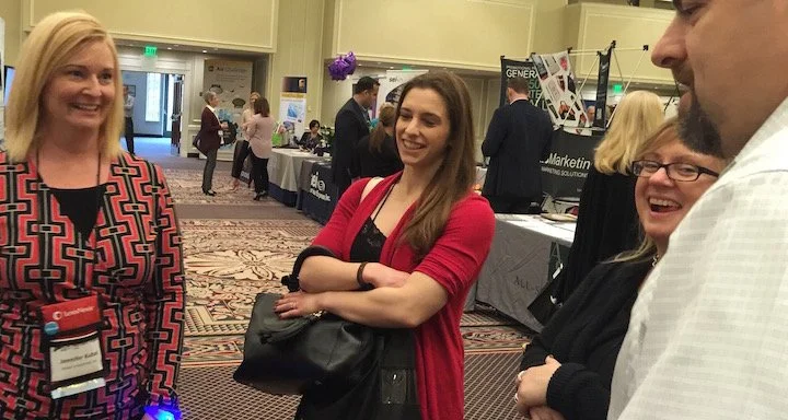 Three women and one man smiling and talking at an indoor conference or trade show, with booths and other attendees in the background.
