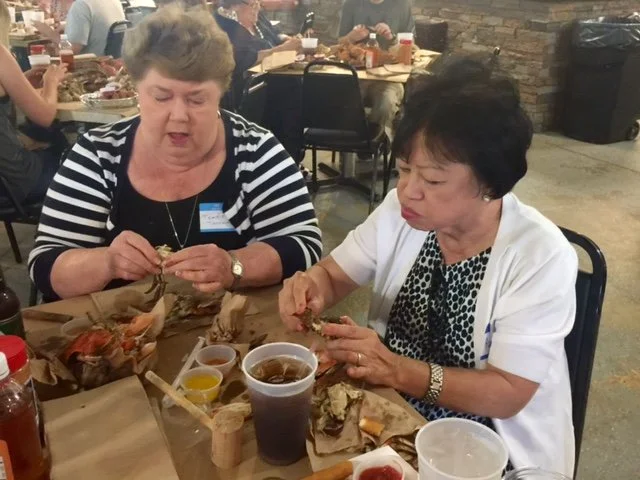 Two women sit at a dining table eating seafood in a restaurant. One woman wears a striped top, and the other wears a white top with a patterned shirt underneath. There are drinks, sauces, and food remnants on the table, and other diners are visible i