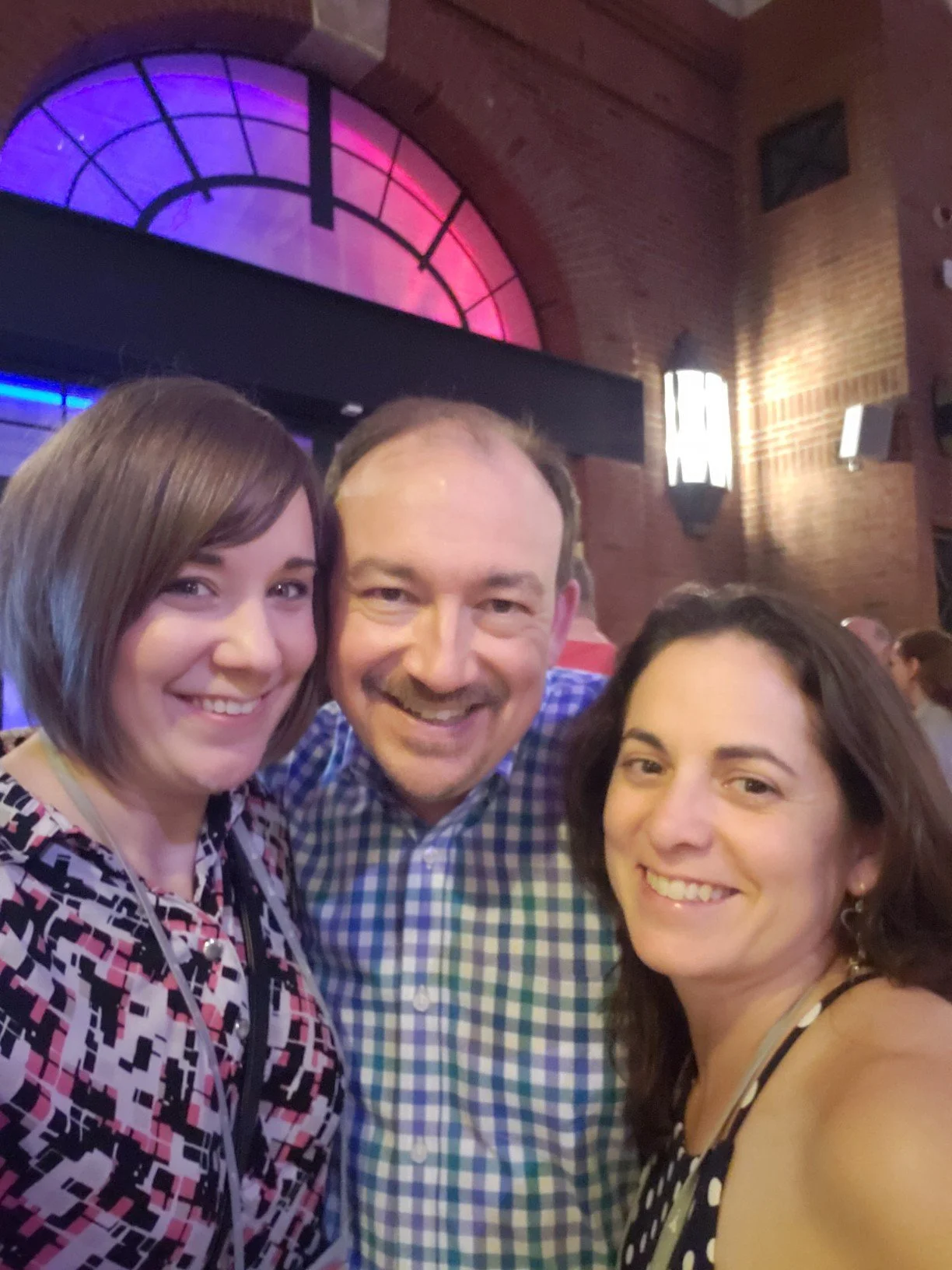 Three people smiling in a dimly lit indoor setting with a circular stained glass window with pink and blue lighting in the background.