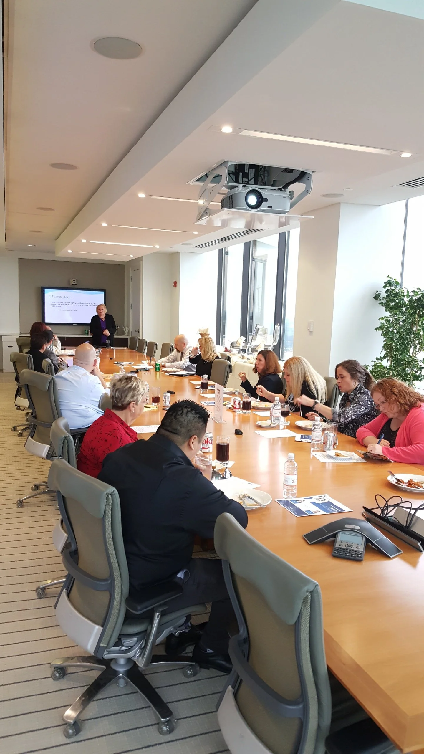 People seated around a long conference table in a modern meeting room, with a presenter at the front, a large screen, and a ceiling-mounted projector.