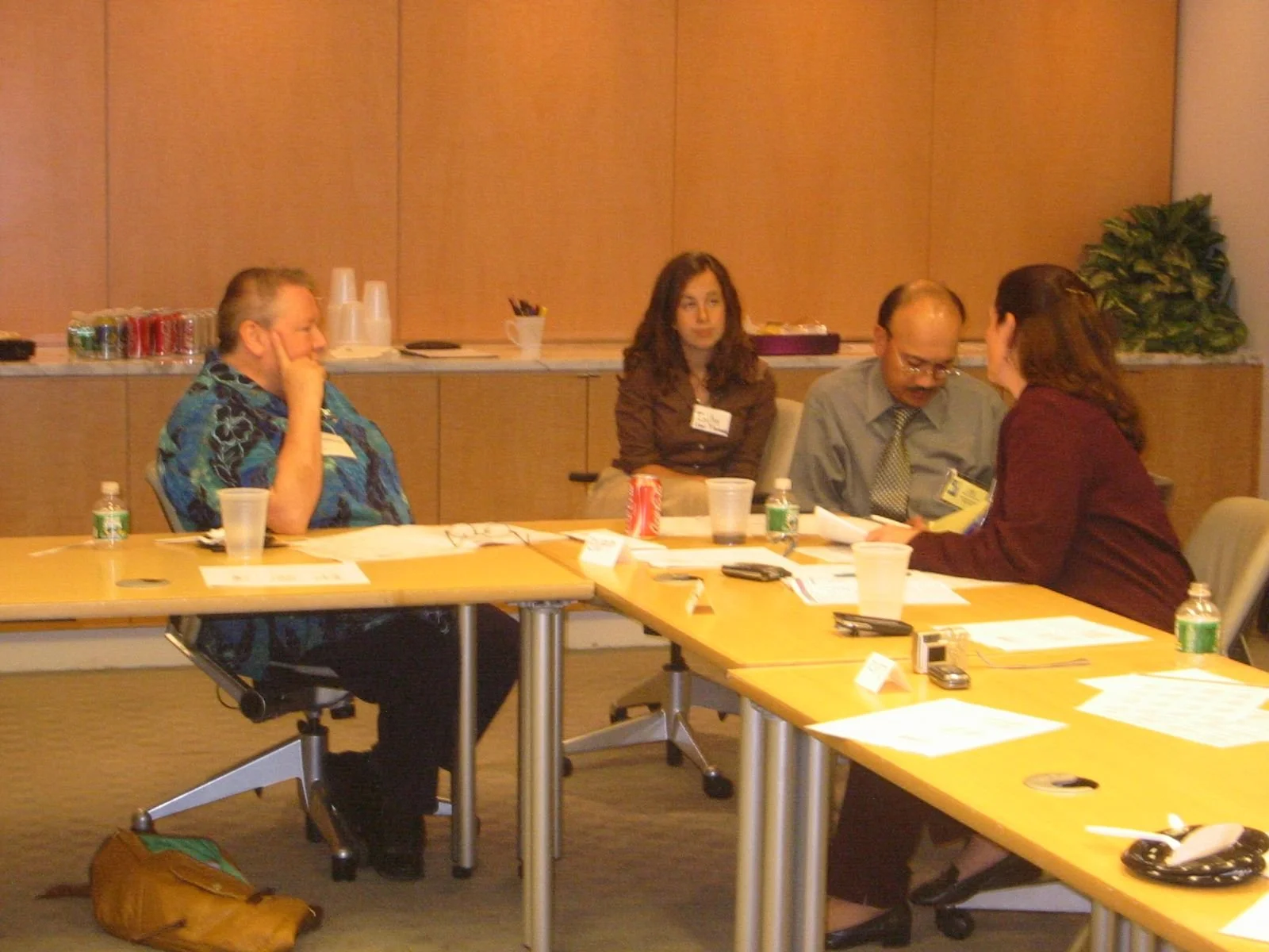 A group of four people sitting at a conference table in a meeting room, with papers, drinks, and electronic devices in front of them.