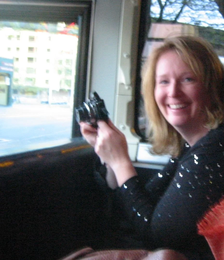 A woman with red hair smiling and holding a black handgun inside a vehicle, with a city street visible through the window.