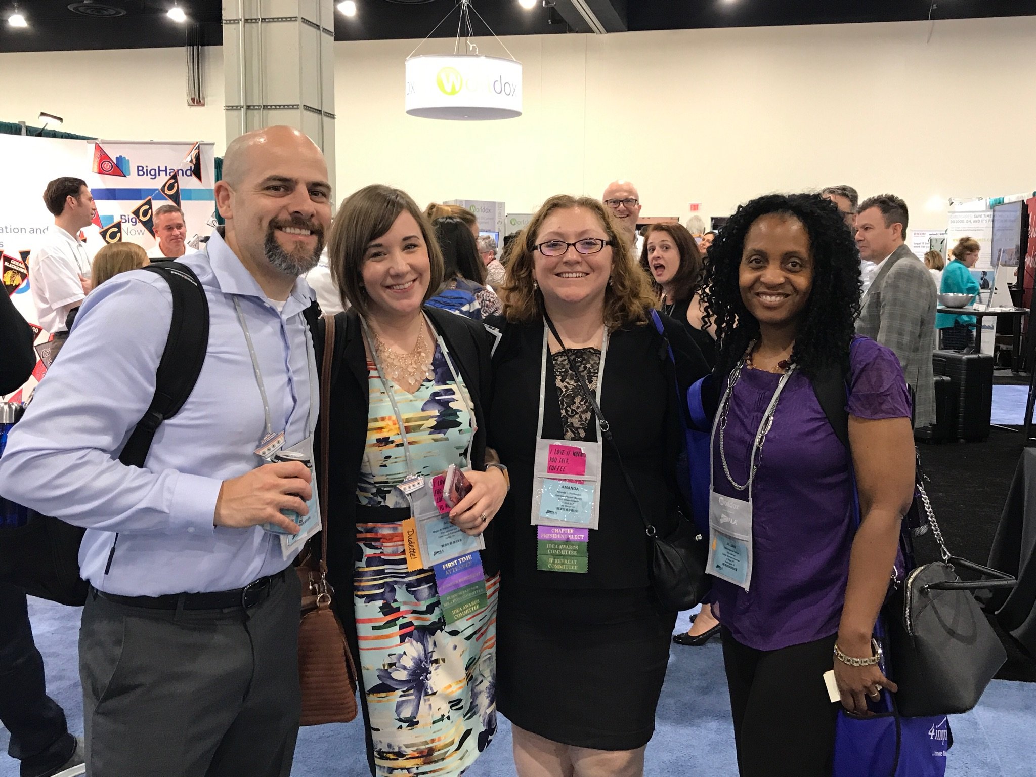 Four smiling women and one smiling man standing together at a conference or trade show, wearing name tags and ribbons, with a crowded background featuring booths and banners.