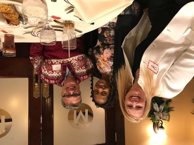 Three women standing together at a dinner table, smiling for the camera. The table has glasses, plates, and napkins, with a floral decoration in the background.