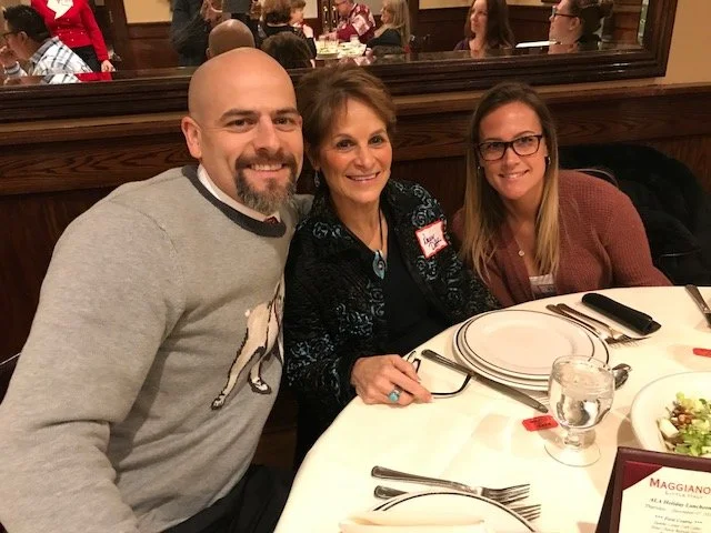 Three people sitting at a restaurant table, smiling at the camera. The table has plates, silverware, a glass of water, and a salad. The background shows other diners and a mirror.