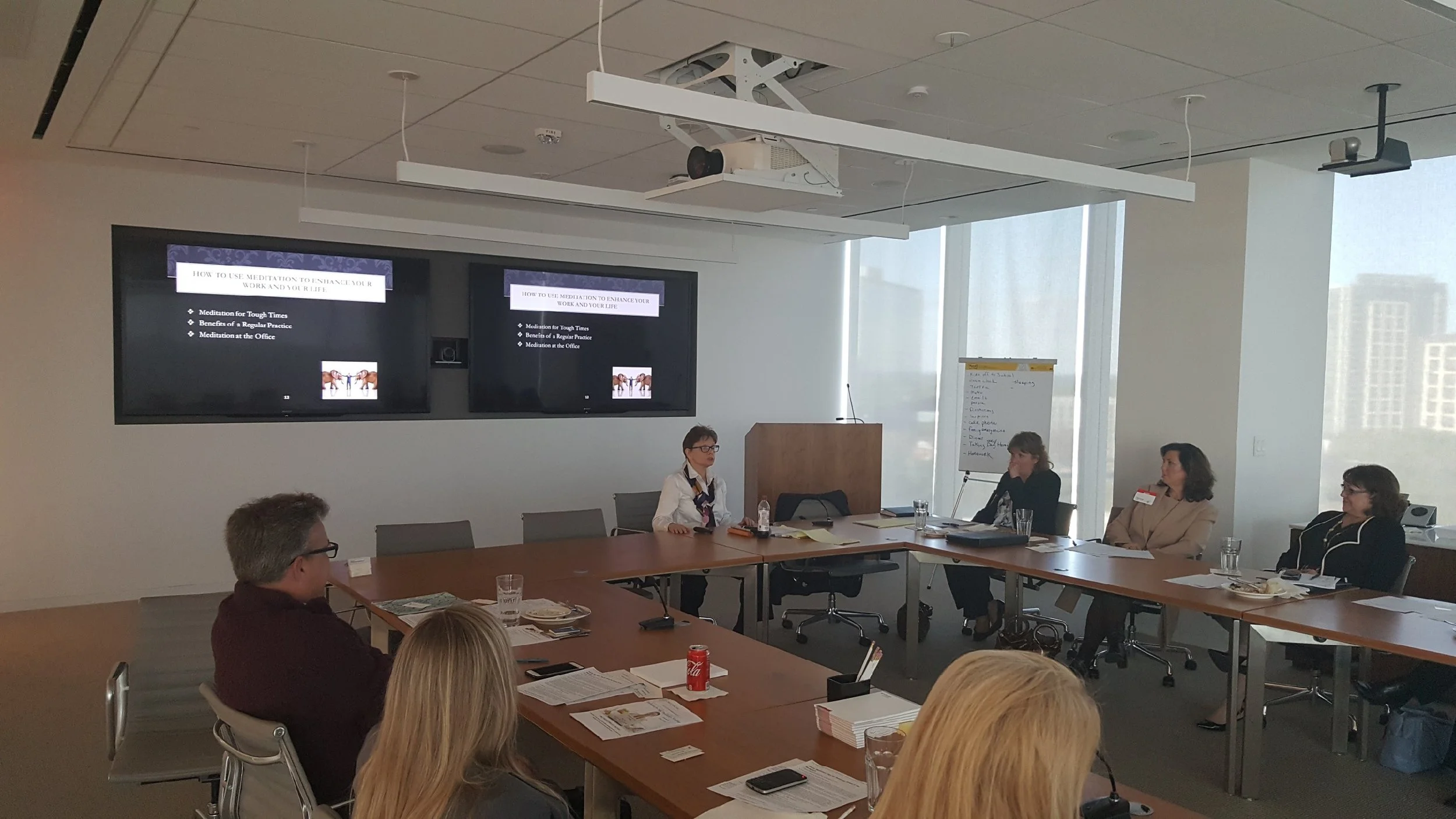 A conference room with a large table and several people seated around it, participating in a meeting. Two large screens display a presentation on meditation and wellness. There are notebooks, papers, water bottles, and a soda can on the table. Light 