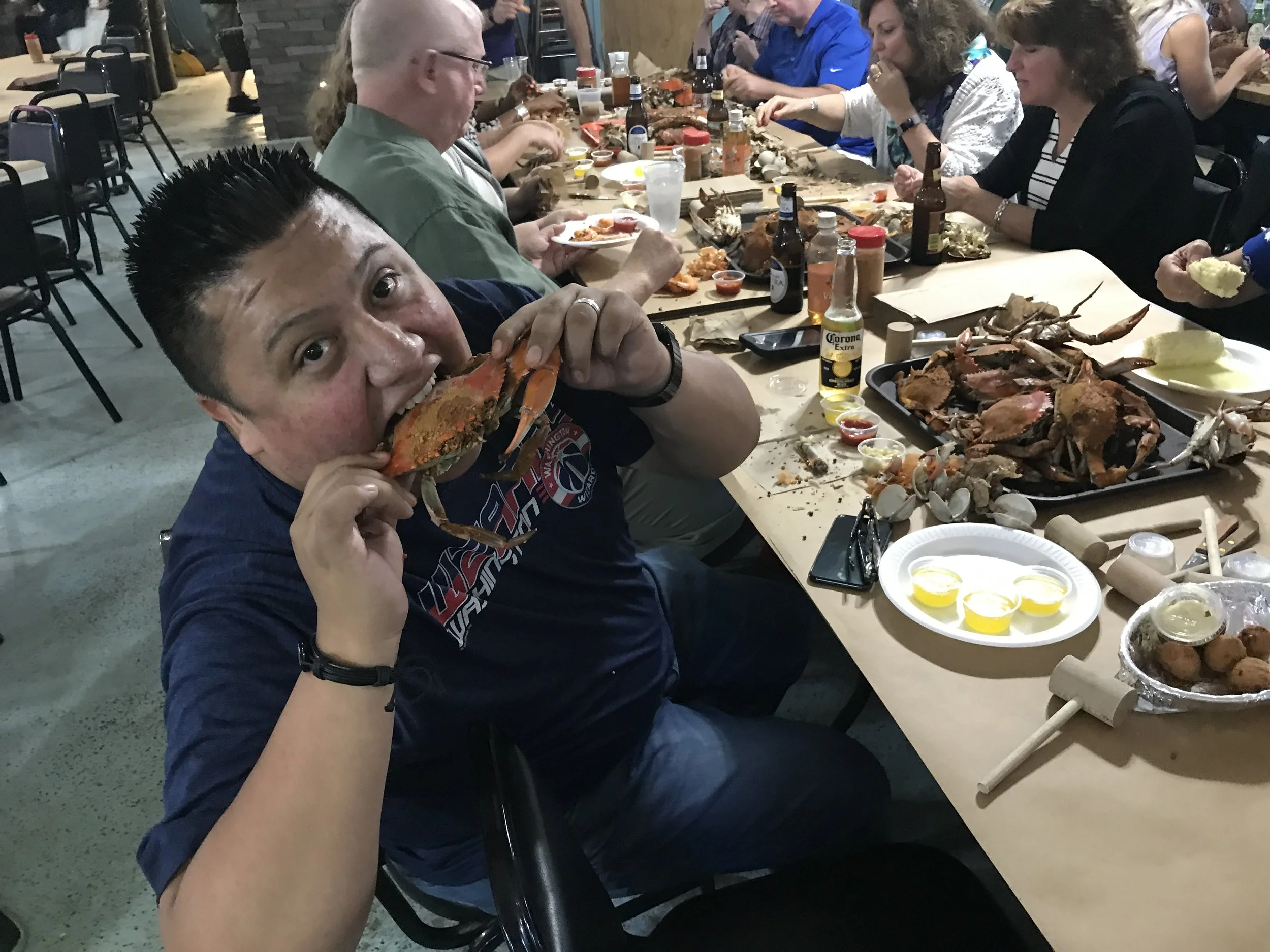 A group of people sitting at a long dining table filled with seafood and side dishes, with one person in the foreground holding a crab and smiling at the camera.