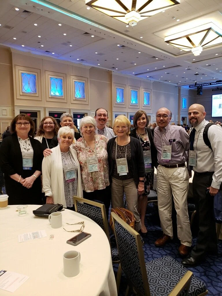 Group of nine professionals at a conference, smiling and posing for a photo in a hotel ballroom with blue decorative lighting.