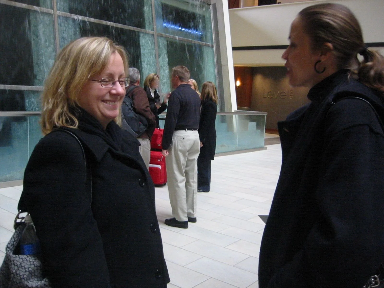 Two women are talking in an indoor setting, with a group of people in the background near a large glass wall or window.