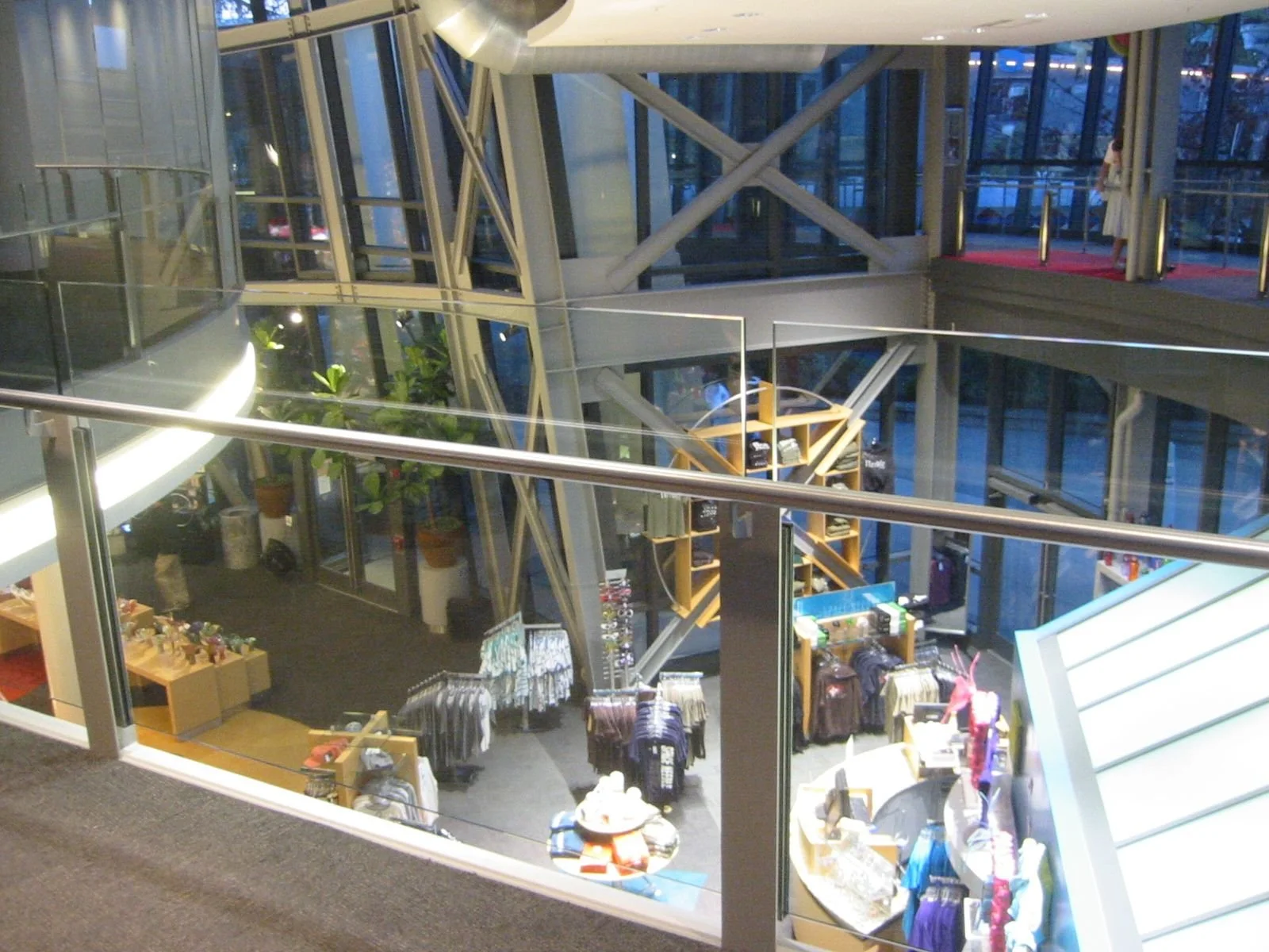 Interior view of a modern shopping mall with merchandise displays on the ground floor and glass walls revealing an upper level with a person walking. The mall features metal beams, glass, and indoor plants.