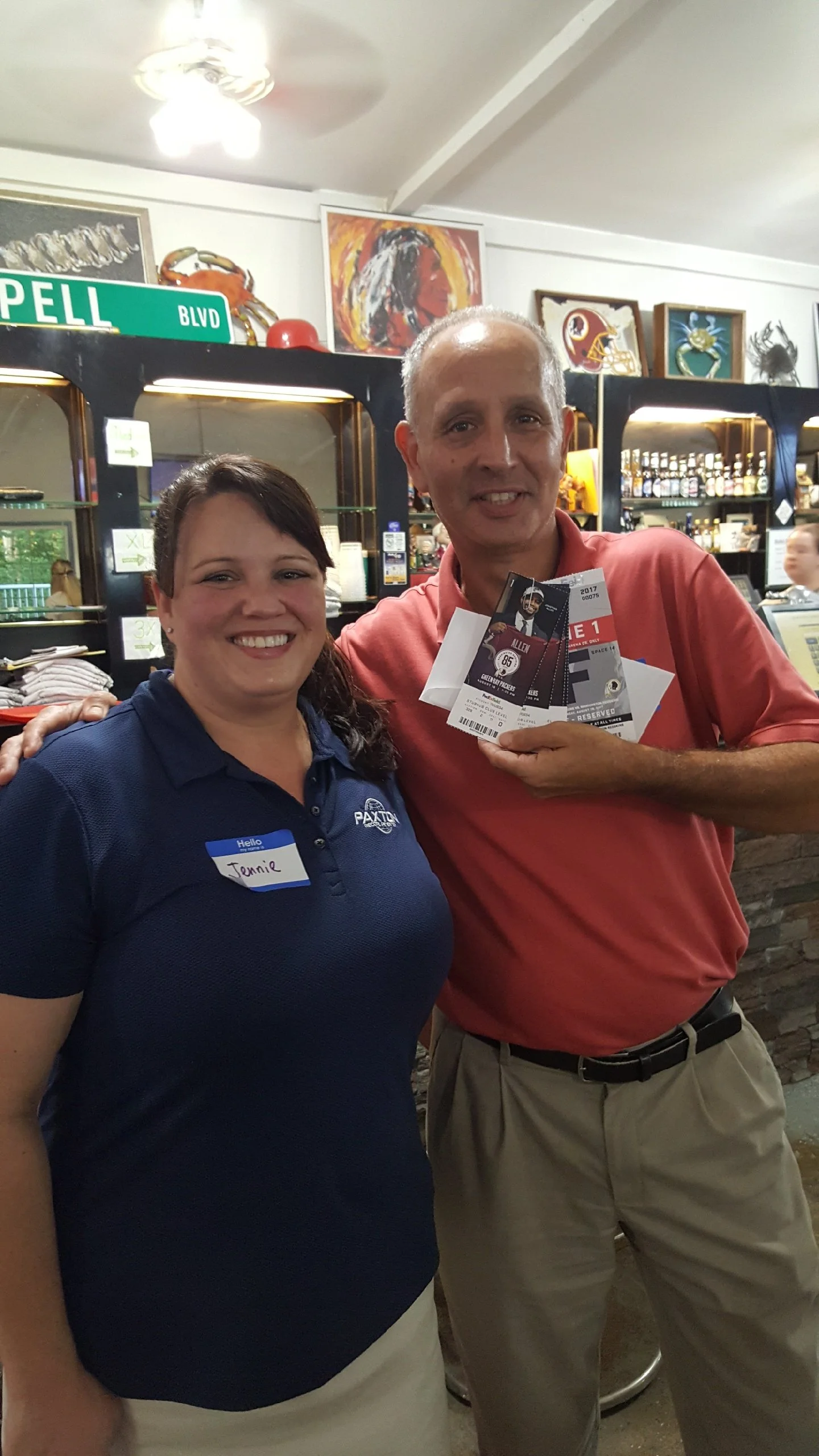 A woman in a blue shirt with a name tag stands next to a man in a pink shirt holding two tickets, in a bar or restaurant setting with artwork and bottles in the background.