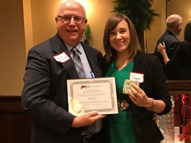 A man and a woman smiling and holding a certificate of achievement. Both are wearing name tags, and the woman is also holding a small item. Other people are visible in the background at a social event.