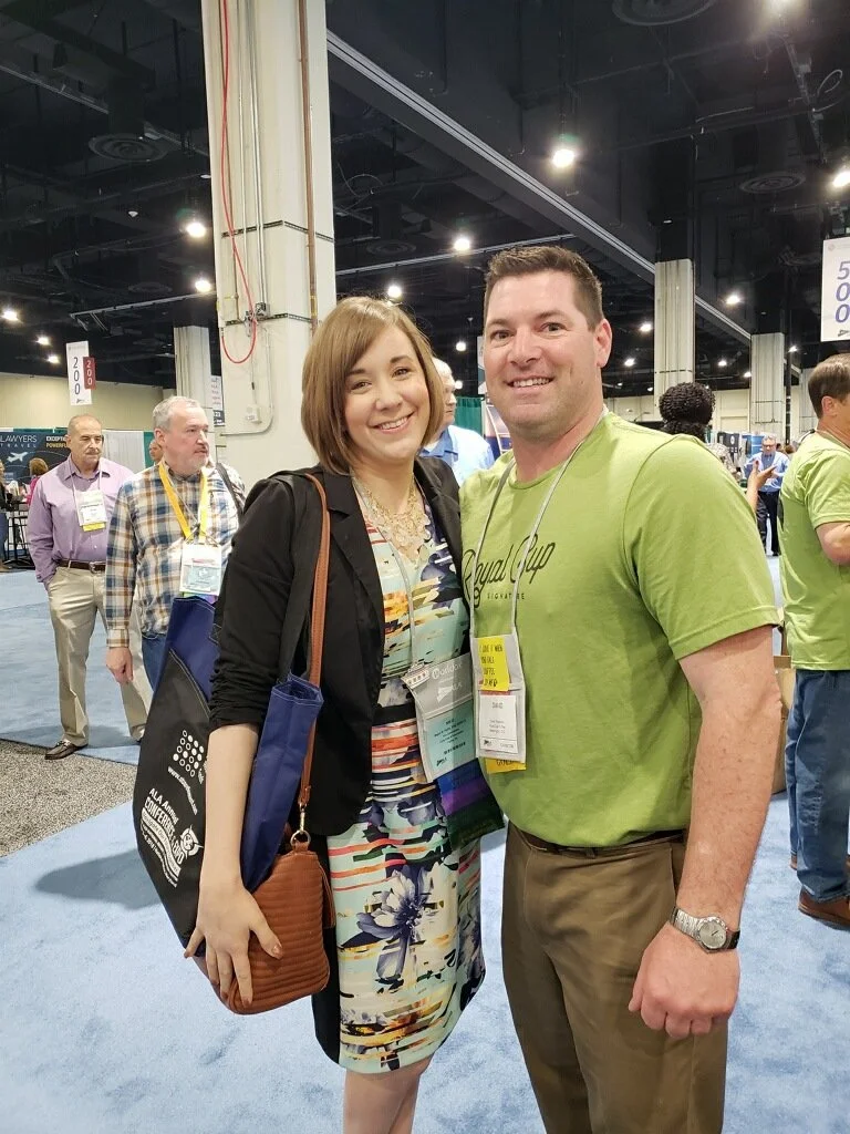 A woman and a man smiling at a conference, with several other attendees and booths in the background.