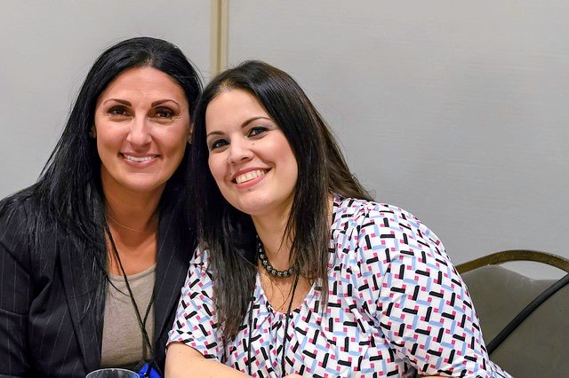 Two women sitting close together, smiling at the camera, in an indoor setting with a plain wall.