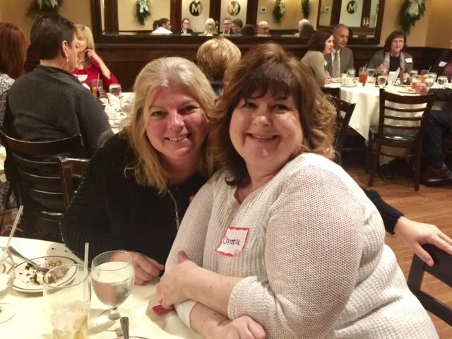 Two women smiling and sitting close together at a restaurant table, with other diners in the background.