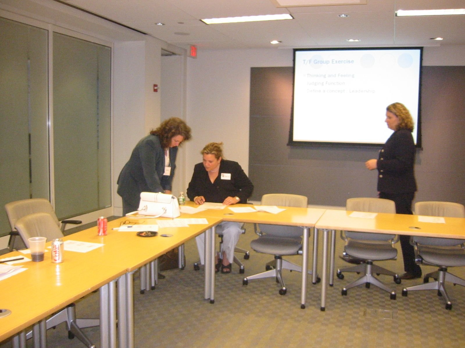 Three women in a conference room with a large yellow table, papers, soda cans, and a projector screen displaying a presentation titled 'T/F Group Exercise.' Two women are standing and one is seated, all engaged with documents.