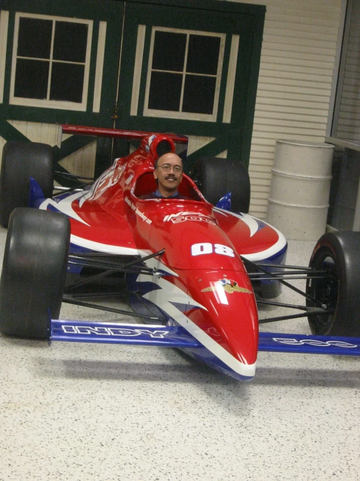 A man with glasses and a mustache sitting in a red, white, and blue vintage formula race car inside a garage area.