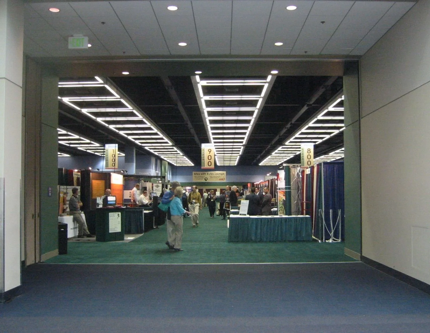 An indoor trade show with multiple booths, visitors walking around, and overhead lighting, entrance seen from outside.