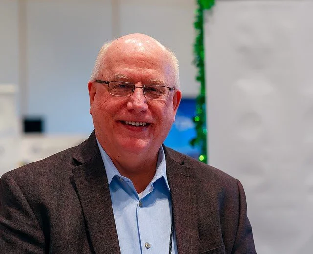 Portrait of a smiling middle-aged man with glasses, wearing a dark suit jacket and light blue shirt, indoors with a blurred background.