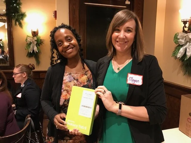 Two women smiling at a gathering, one holding a yellow Samsung Galaxy Tab E box, the other wearing a name tag that says 'Megan', surrounded by decorated walls with holiday wreaths.