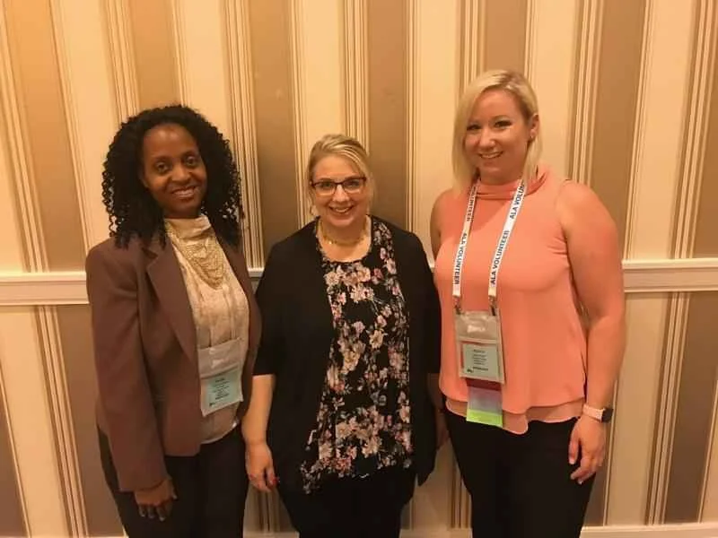 Three women standing together and smiling at a conference or event, with beige striped wallpaper in the background.