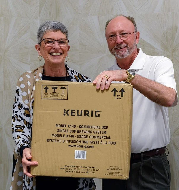A smiling older woman holding a box with a man standing beside her, both standing in front of a beige wall. The woman wears glasses and a patterned scarf, while the man wears glasses and a white polo shirt. The box they hold is labeled "KEURIG" and a