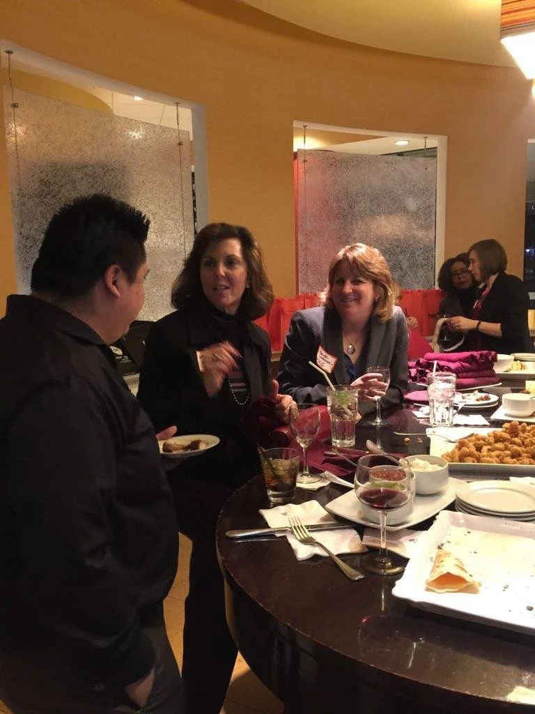 People gathered around a table at a restaurant or banquet, enjoying drinks and food, engaged in conversation, with desserts and dishes on the table.