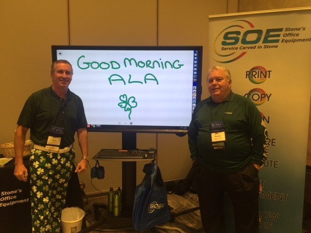 Two men standing next to a digital whiteboard with a message that says 'Good Morning ALA' and a shamrock drawing. One man is wearing a dark shirt and floral pants, the other is wearing a dark shirt and dark pants. They are in a conference or event se