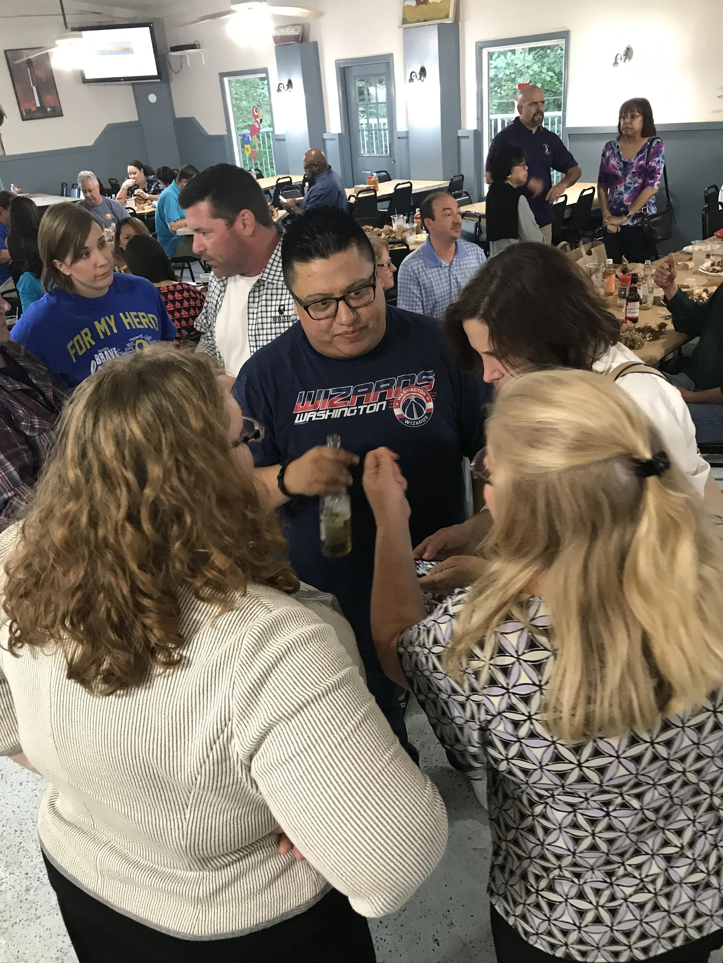 People gathered in a room, engaged in conversations, with several tables and windows with greenery outside. Some individuals wear casual attire, and the atmosphere appears social.