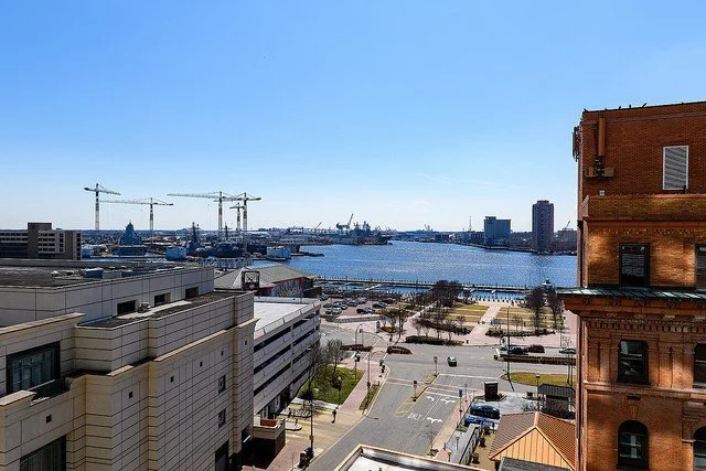 View of a harbor with boats, cranes, and a city skyline in the background, with streets and buildings in the foreground.